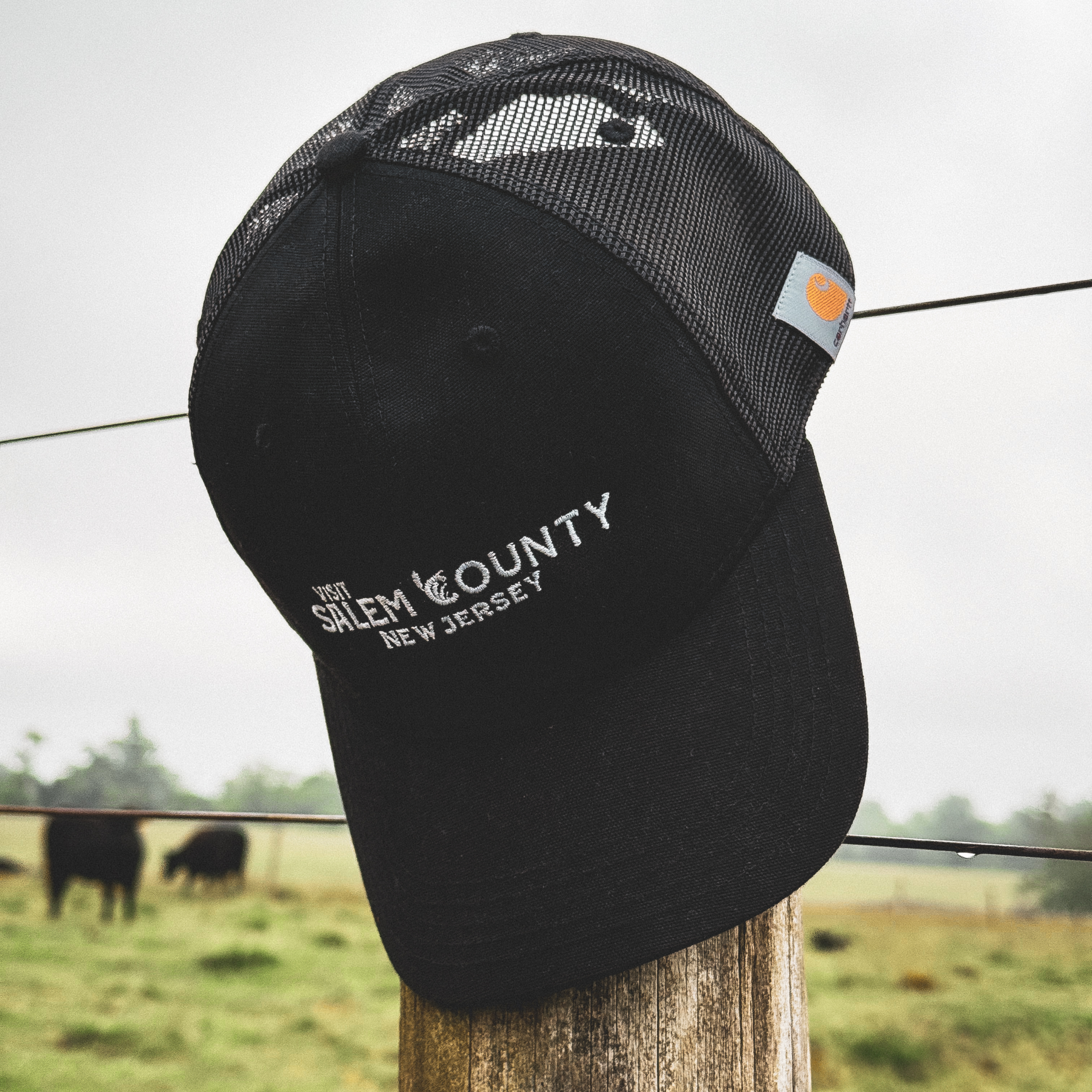 A black Visit Salem County New Jersey logoed baseball cap hanging from a fence post in the country.